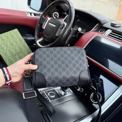 A grey monogrammed crossbody bag held up inside a car, above the centre console and gear selector