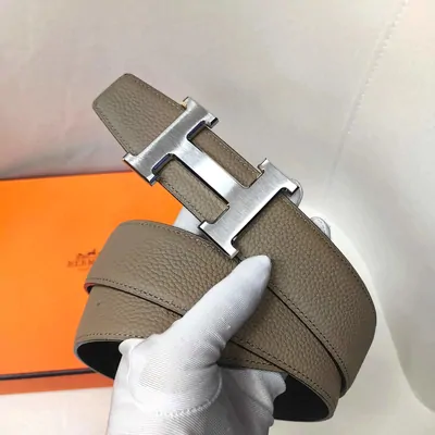A taupe leather belt with a wide silver buckle, held in a white-gloved hand beside its orange box