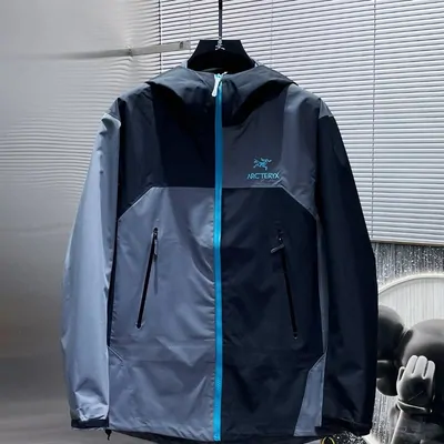 A navy and slate blue hooded shell jacket hanging zipped shut in front of a window blind