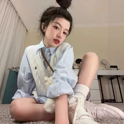 A model sitting on a rug in a blue striped shirt under a white cable knit sweater vest, with white socks and trainers